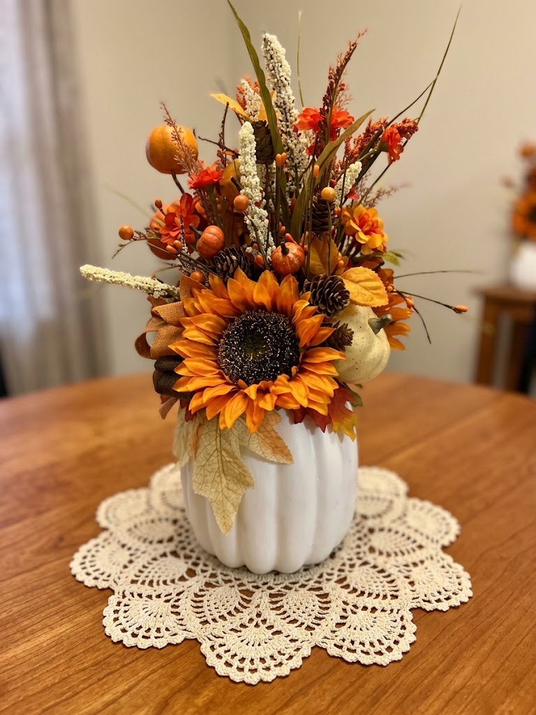 Handmade fall floral arrangement in a white ribbed pumpkin vase with sunflower, orange berries, pine cones, and autumn botanicals on a lace doily